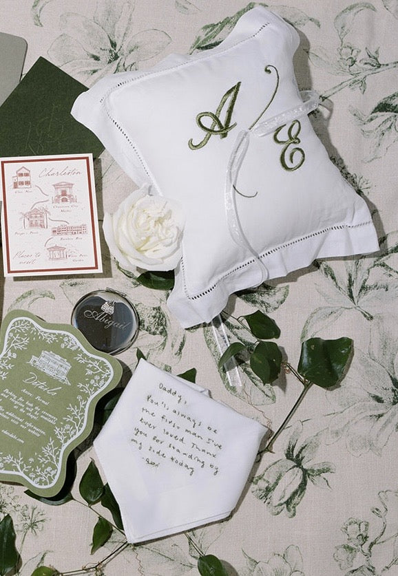 White embroidered ring pillow with monogram, card, and handkerchief on floral background