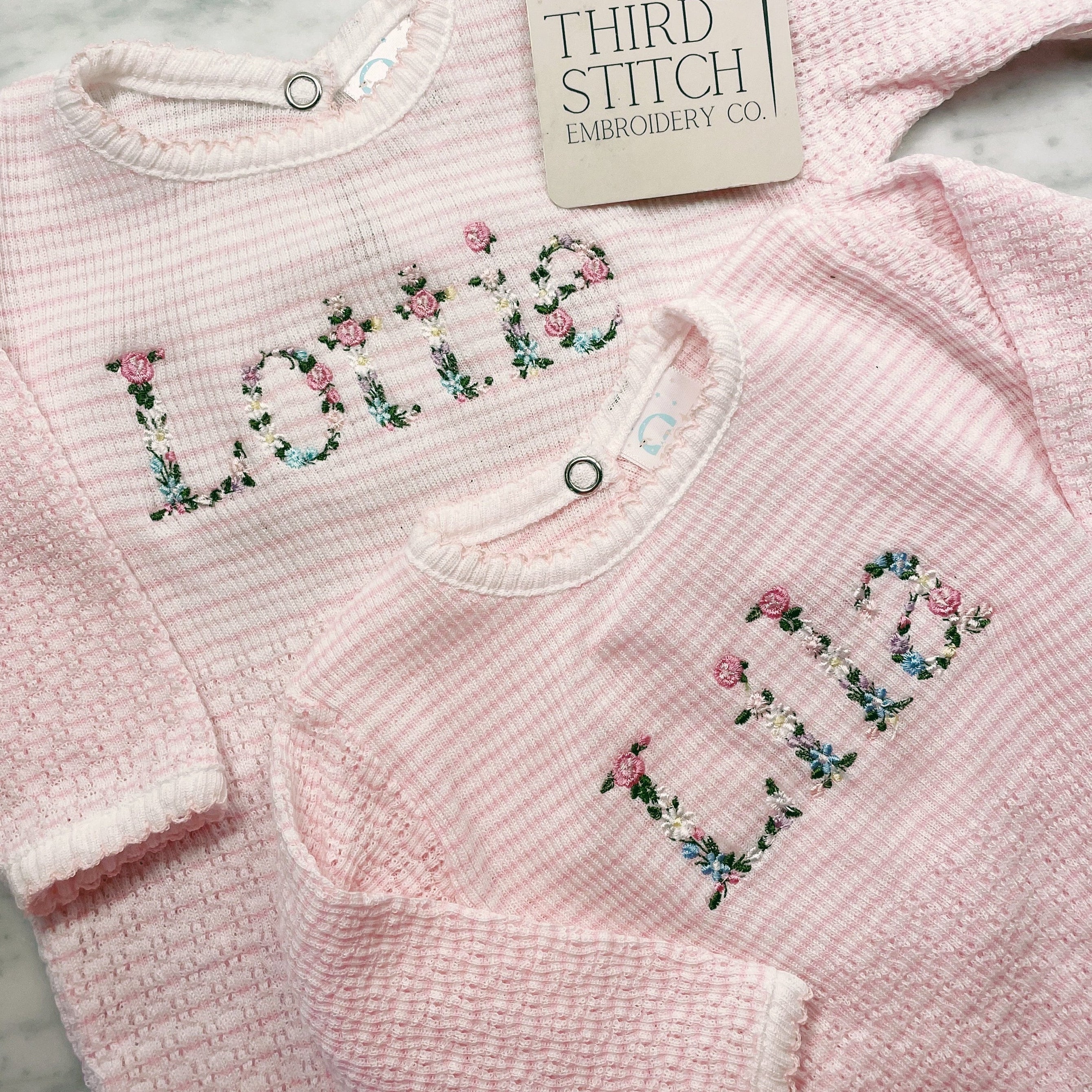 Pink baby sweater with floral text embroidery on a textured surface