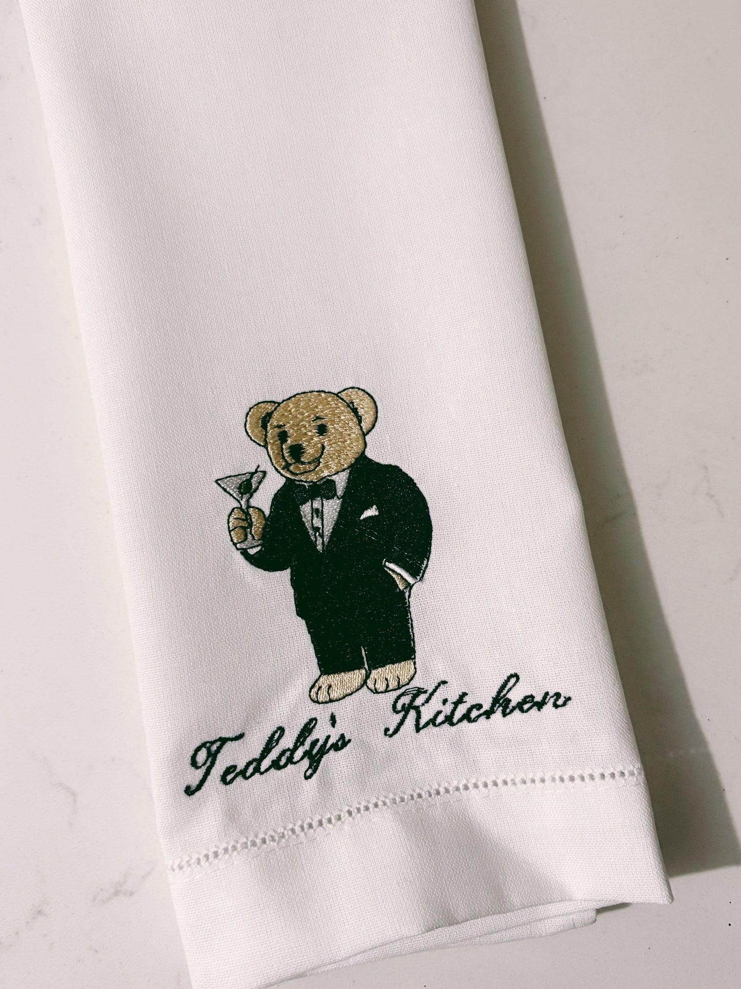 White towel with embroidered teddy bear in a tuxedo and 'Teddy's Kitchen' text on a light gray background