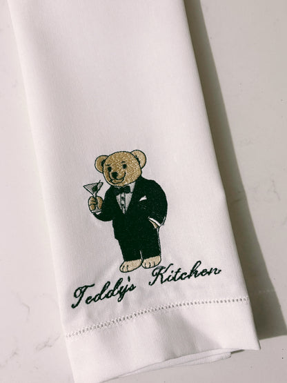 White towel with embroidered teddy bear in a tuxedo and 'Teddy's Kitchen' text on a light gray background